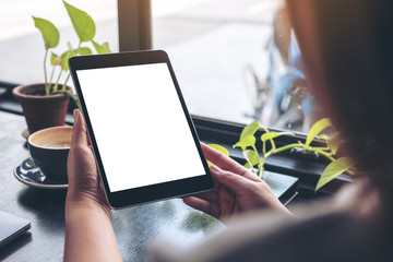 Mockup image of a woman holding black tablet pc with blank white desktop screen with green leaves and coffee on wooden table