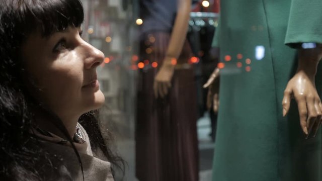 Outdoor portrait of beautiful woman in coat looking at the shop window. Closeup of young woman looking at boutique showcase in the evening city on the street.