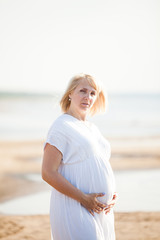 Pregnant woman in white dress posing on the beach at sunny evening. High key portrait