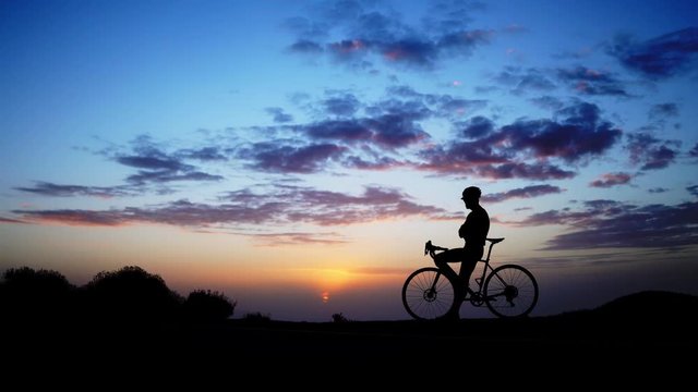 A man on a Bicycle stands on top of the mountain and admires the sunset. The camera moves on a Steadicam.