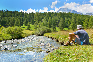 hiker sitting at the edge of a river in the feet of alpine mountains