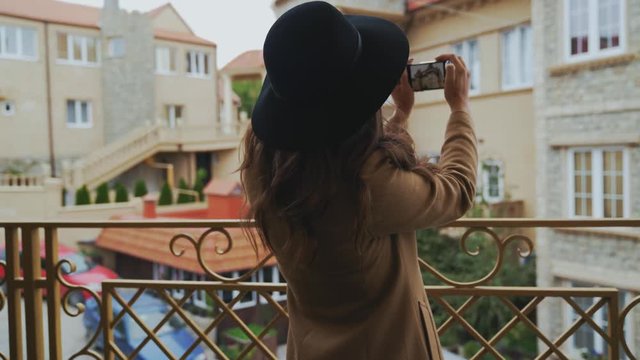 View from the back, close-up, steadikam slender woman in a coat and hat walks around the city, takes a photo of the architect on her smartphone, enjoys a walk. The girl flew over the weekend to Europe