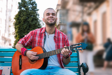 Life is music. Low angle of attractive street musician having seat on bench while strumming strings...