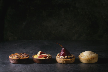 Variety of sweet tartlets with chocolate, caramel, pears, figs in row on black texture table.