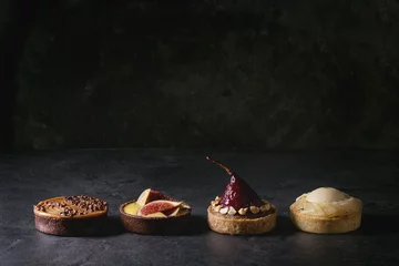 Poster Variety of sweet tartlets with chocolate, caramel, pears, figs in row on black texture table. © Natasha Breen