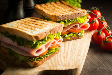 Close-up of two sandwiches with bacon, salami, prosciutto and fresh vegetables on rustic wooden cutting board. Club sandwich concept.