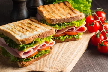 Foto op Plexiglas Snackbar Close-up van twee sandwiches met spek, salami, prosciutto en verse groenten op rustieke houten snijplank. Clubsandwich-concept.