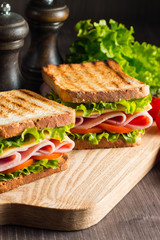 Close-up of two sandwiches with bacon, salami, prosciutto and fresh vegetables on rustic wooden cutting board. Club sandwich concept.