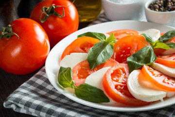 Photo of Caprese Salad with tomatoes, basil, mozzarella, olives and olive oil on wooden background. Italian traditional caprese salad ingredients. Mediterranean, organic and natural food concept.