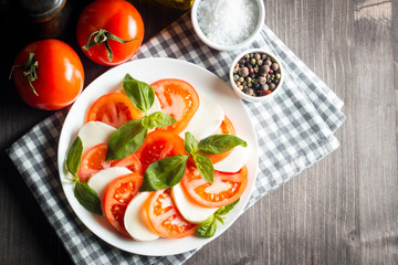 Photo of Caprese Salad with tomatoes, basil, mozzarella, olives and olive oil on wooden background. Italian traditional caprese salad ingredients. Mediterranean, organic and natural food concept.
