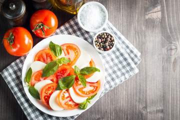 Photo of Caprese Salad with tomatoes, basil, mozzarella, olives and olive oil on wooden background. Italian traditional caprese salad ingredients. Mediterranean, organic and natural food concept.