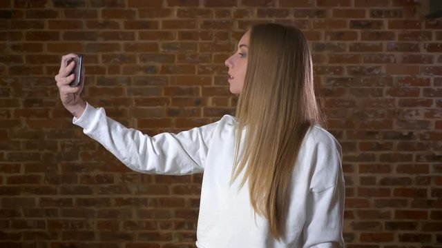 Good-looking caucasian blonde woman is taking pictures on web camera of her phone, standing relaxed in profile in brick studio alone