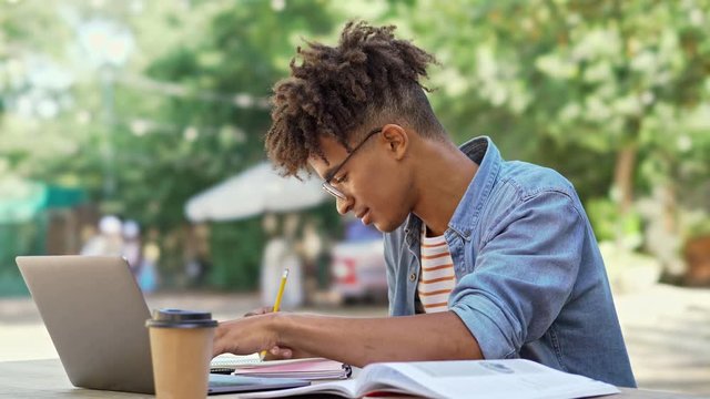 Smiling african student in eyeglasses using laptop computer and writing somthing while sitting by the table in cafe outdoors