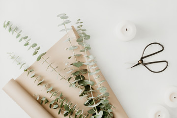 Creating of a bouquet with baby blue eucalyptus branch in a golden wrapping paper among white candles on a table. The concept of a florist work or celebration. Top view, flat lay. - obrazy, fototapety, plakaty