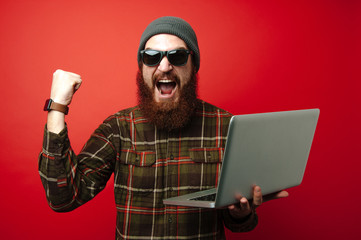Cheerful bearded hipster man using laptop and celebrating success with amazed face expression