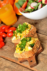 Close-up of wholegrain slices of bread with scrambled eggs, fresh herbs, spring onions, tomatoes, orange juice and salad bowl in background - healthy snack