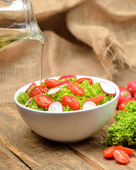 Pouring oil into green salad Lollo Biondo with tomatoes and radishes in a white bowl on wooden table, a pitcher of oil in the background - vertical photo