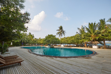 Swimming pool side of luxury hotel ith deck chairs, palm trees and blue ocean. Maldives