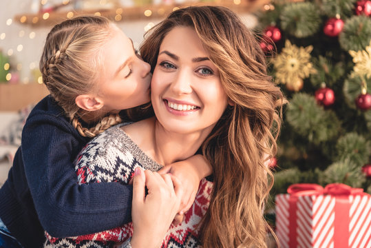 adorable daughter kissing smiling mother near christmas tree at home