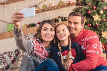 Naklejka premium parents and daughter taking selfie with smartphone near christmas tree at home