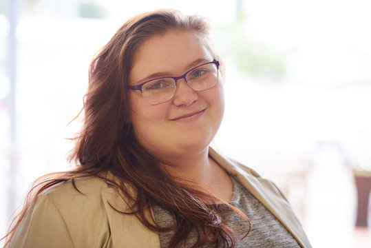 close up portrait of beautiful plus size young adult woman, with brown hair and a beige blazer, while looking into the camera with her prescription glasses on.