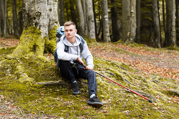 Young blonde attractive smiling man with trekking poles and camera sitting and relaxing on the moss in the forest, trekking, recreation and healthy lifestyle concept