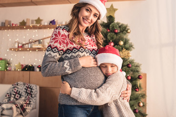 daughter in santa hat hugging pregnant smiling mother near christmas tree at home