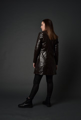 full length portrait of brunette woman wearing long leather coat. standing pose  with back to the camera, on grey studio background.