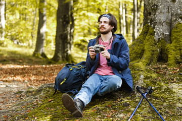 Horizontal side view portrait of caucasian young brunette bearded man wearing pink t-shirt, blue cloak and jeans holding camera and meditating relaxing alone on moss front of tree surrounded by forest