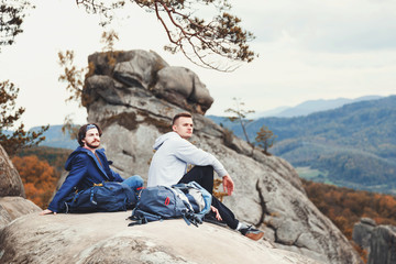 Two young hipster travellers with backpacks sitting on the rocks and looking on the forest,...