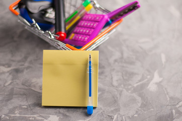 Blank paper sticker and blue ball pen near full metal market basket of school supplies on gray concrete background with copy space for your text. Education or business concept