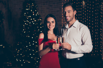 Portrait of attractive, handsome man in formal wear and lady in trendy, stylish red dress hold cocktail, look at camera stand in room with lights garland decorations on pine tree