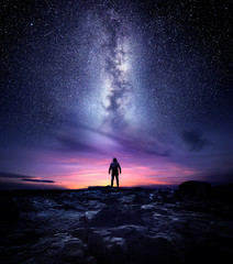 Night time long exposure landscape photography. A man standing in a high place looking up in wonder to the Milky Way galaxy, photo composite. - obrazy, fototapety, plakaty