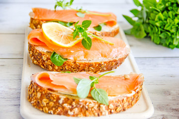  Sandwices with salted salmon and soft white bread cheese, decorated with a slice of lemon and fresh greens on an wooden background. Culinary background with appetizer of bread and red fish