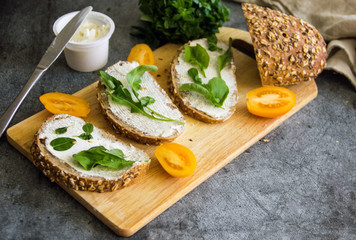 Three sandwiches with cheese lie in a row on a white board on a dark background, decorated with greens, next to cheese in a jar, sprigs of greens and fresh tomatoes, top view
