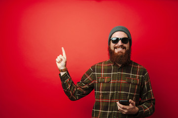 Young happy bearded man holding phone and pointing up at copyspace