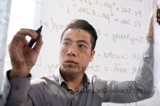 Shot through glass board of concentrated Asian man writing code on it 