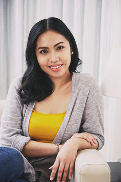 Gorgeous Filipino female smiling and looking at camera while relaxing on comfortable sofa in cozy living room