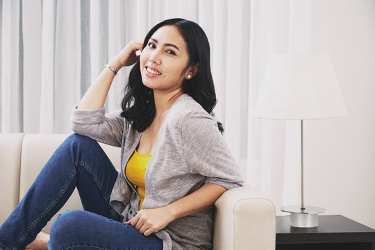 Side view of attractive Filipino female smiling and looking at camera while sitting on soft couch near lamp in stylish living room