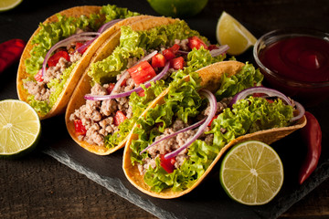 Photo of Mexican tacos with ground meat, beef, beans, onions and salsa on wooden background. Ketchup sauce and lime. A glass o beer in the background.