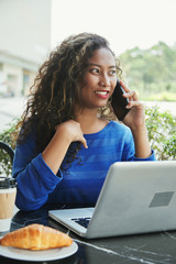 Pretty Indonesian businesswoman smiling and looking away while having smartphone conversation in nice outdoor cafe