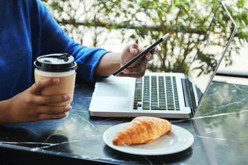 Anonymous Indonesian female holding cup of hot drink and using modern smartphone while sitting at table in outdoors cafe