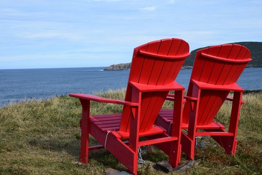 closeup of two empty red Adirondack chairs on the edge of a cliff overlooking the ocean and coastline, Silver Mine Head Path Torbay Newfoundland Canada  