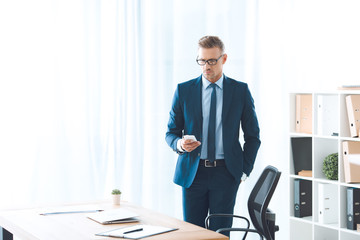 handsome businessman in eyeglasses and formal wear using smartphone at workplace