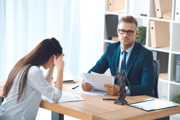 Fototapeta premium lawyer holding papers and looking at camera while sad female client sitting in office
