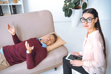 psychotherapist in eyeglasses using digital tablet and looking at camera while patient lying on couch