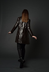 Fototapeta premium full length portrait of brunette woman wearing long leather coat. standing pose with back to the camera, on grey studio background.