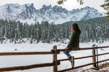 A young woman is sitting on the background of a winter lake and mountains. The girl admires the picturesque winter landscape: a snow-covered lake and mountains. Winter mountain in Italy, Carezza Lake