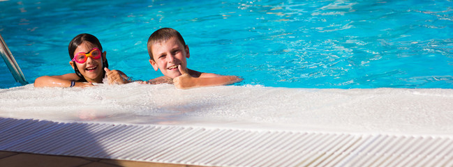 Smiling boy and girl with pink goggles with thumbs up by swimming pool edge. Little kids enjoying...