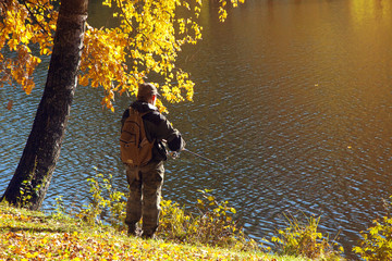 A fisherman with a fishing rod standing on the shore of a lake in sunny autumn weather and birch with yellow  leaves